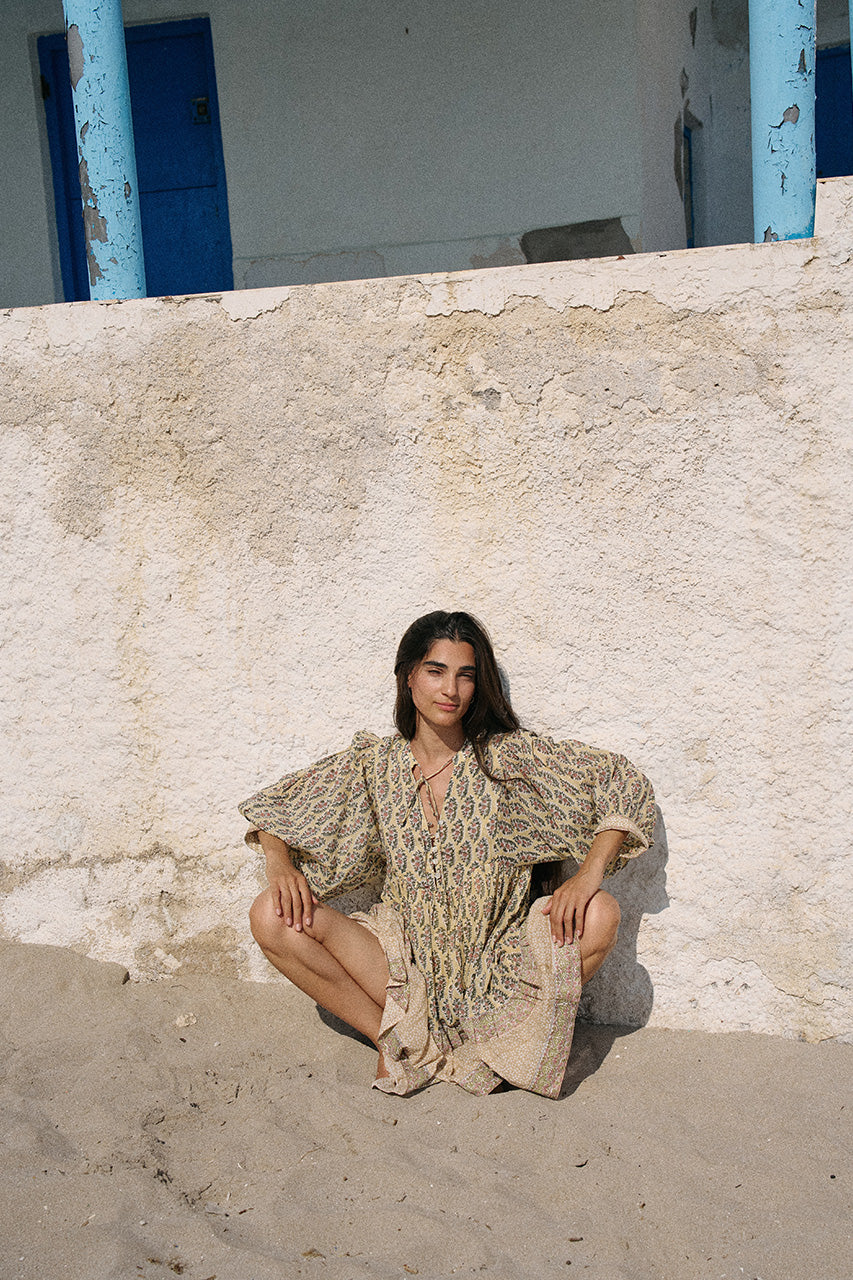 Woman seated on sandy ground against a rustic white wall with blue shutters wearing the Daughters of India Bhoomi Mini Dress in Meadow