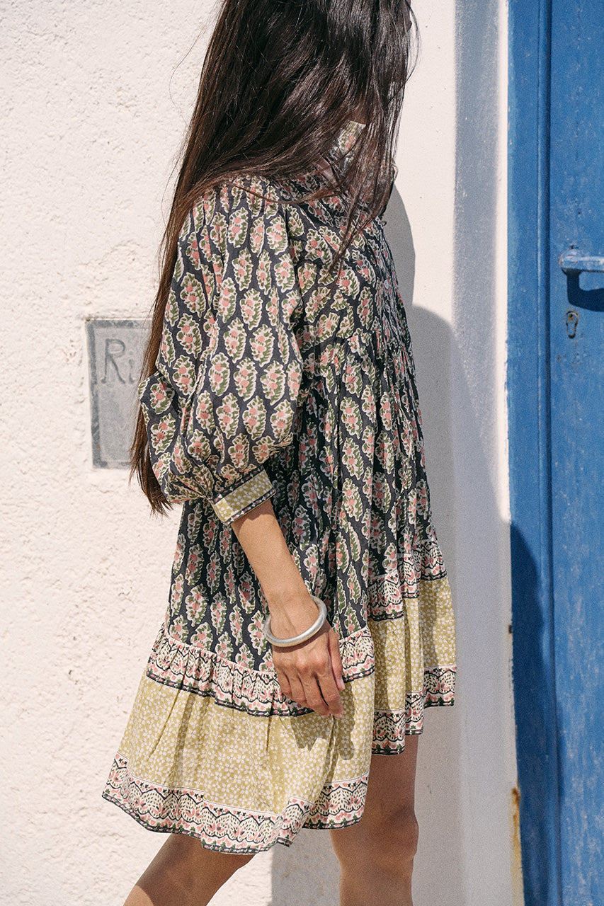 Back view of woman beside a blue door wearing the Daughters of India Bhoomi Mini Dress in Onyx, showing the tiered hem and intricate border print detail at the base of the skirt
