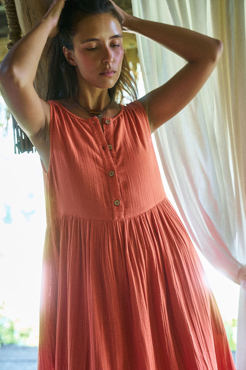 Nadi Dress Terracotta — three-quarter view hands in hair beside sheer white curtain, terracotta double gauze cotton midi dress with wooden button front and gathered skirt, soft natural light | Daughters of India