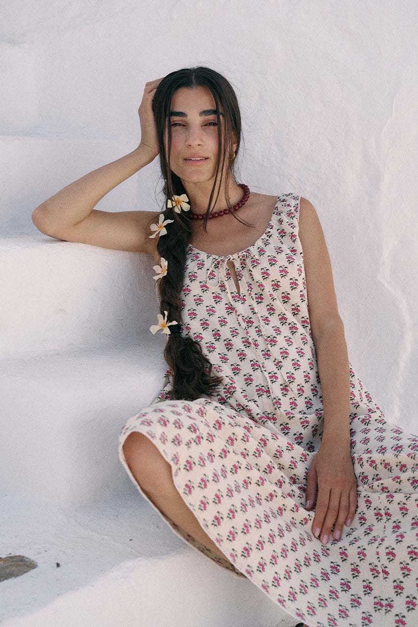 Woman seated on white stone steps in Greece wearing the Daughters of India Sundress Midi in Peony, with floral hair accessories and layered necklace
